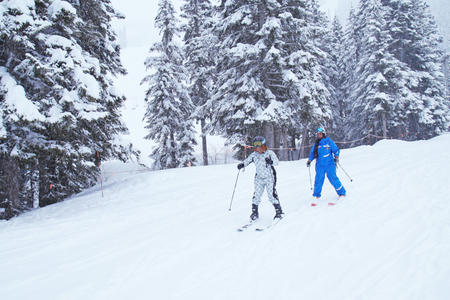 Black woman in white snowsuit and black woman in blue snowsuit ski down snowy slope surrounded by snow filled pine trees